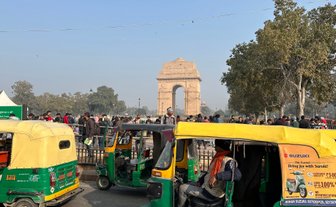 Bunte Tuk-Tuks stehen in der Nähe des India Gate, umgeben von einer Menschenmenge und Bäumen unter klarem Himmel.