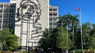 Ein rotes Cabrio fährt vorbei an einem großen Wandbild von Che Guevara, umgeben von Palmen und einem blauen Himmel.