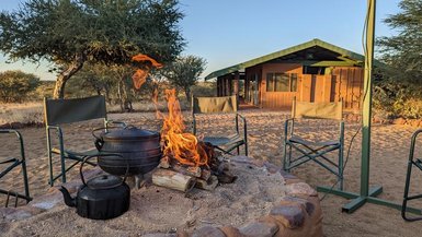 Ein gemütliches Lagerfeuer brennt in einem Steinkreis, umgeben von Campingstühlen und einer rustikalen Unterkunft im Hintergrund.