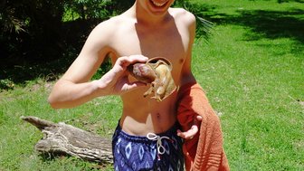 Ein junger Mann hält eine große Schnecke in der Hand und lächelt, während er auf einer grünen Wiese steht.
