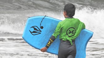 Ein Junge in einem grünen Neoprenanzug trägt ein blaues Surfbrett und blickt auf die Wellen des Ozeans.