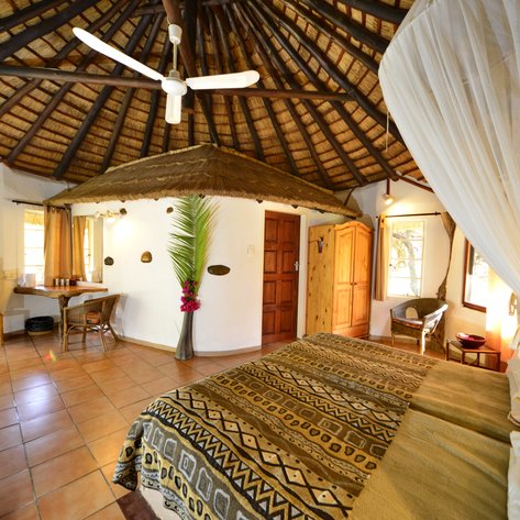 Ein gemütliches Zimmer mit einem Strohdach, einem großen Bett und einem Tisch, umgeben von warmen Erdtönen und natürlichem Licht.