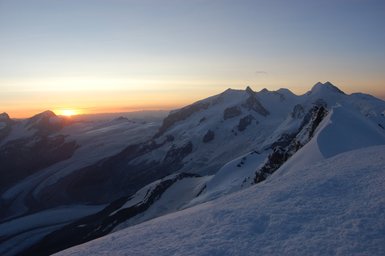 Die schneebedeckten Berge erstrahlen im warmen Licht der untergehenden Sonne, während der Himmel in sanften Farben leuchtet.