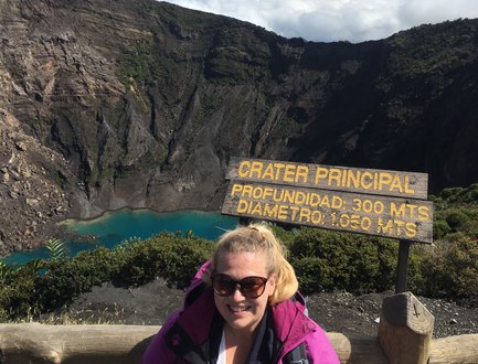 Eine Frau mit Sonnenbrille steht vor einem beeindruckenden Vulkan-Krater mit türkisfarbenem Wasser und einer Holztafel.