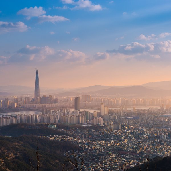 Die Skyline von Seoul bei Sonnenuntergang, mit dem markanten Lotte World Tower, umgeben von sanften Hügeln und Wolken.