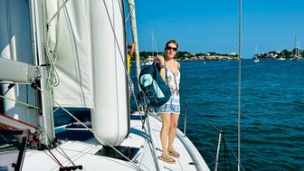 Eine Frau steht auf dem Deck eines Segelbootes, während sie eine Tasche trägt und den klaren blauen Himmel betrachtet.