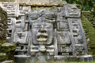 Ein beeindruckendes, verwittertes Steinrelief mit einem menschlichen Gesicht und mysteriösen Symbolen, umgeben von üppigem Grün.
