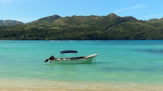Ein kleines Boot mit einem blauen Sonnendach schwimmt ruhig im klaren, türkisfarbenen Wasser vor einer grünen Berglandschaft.