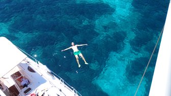 Ein Mann schwimmt entspannt im klaren, blauen Wasser, während ein Boot im Hintergrund zu sehen ist.