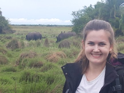 Eine lächelnde Frau steht in einer grünen Landschaft, während im Hintergrund zwei Nashörner grasen.