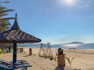 Ein sonniger Strand mit Palmen, Liegen und einem Strohdach, das Schatten bietet, während das Wasser sanft plätschert.
