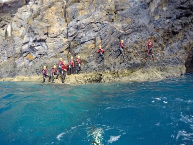 Eine Gruppe von Abenteurern in Neoprenanzügen klettert an einer steilen Felswand entlang, umgeben von klarem, blauem Wasser.