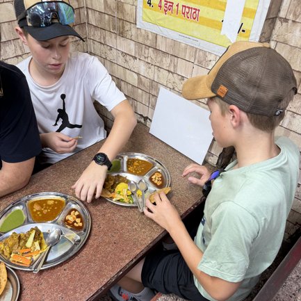Zwei Jungen sitzen an einem Tisch und genießen indisches Streetfood von einem runden Metalltablett mit verschiedenen Gerichten.