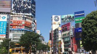 Eine belebte Straßenszene in Tokio mit Menschen, die unter bunten Werbetafeln und strahlend blauem Himmel gehen.