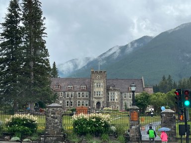 Ein historisches Gebäude mit steiniger Fassade steht vor einer bewaldeten Berglandschaft, umgeben von grünen Bäumen.