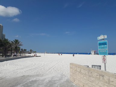 Ein sonniger Strand mit feinem, weißem Sand und blauen Sonnenschirmen, die in der Ferne aufgestellt sind.