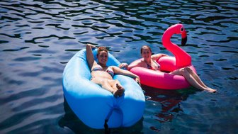 Zwei Frauen entspannen auf bunten Luftmatratzen im klaren Wasser, umgeben von glitzernden Wellen und Sonnenschein.