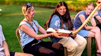 Eine Frau mit Zöpfen lächelt, während sie ein traditionelles Musikinstrument in einer Gruppe von Menschen hält.