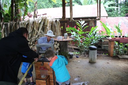 Ein Mann zeigt Kindern, wie Zuckerrohr mit einer Presse verarbeitet wird, um Saft zu gewinnen, umgeben von tropischer Vegetation.