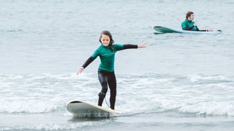 Ein Mädchen surft fröhlich auf einem Surfbrett, während ein anderer Surfer im Hintergrund im Wasser sitzt.