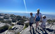 Drei Kinder stehen auf einem Felsen und blicken auf eine weite Landschaft mit Meer und Bergen im Hintergrund.