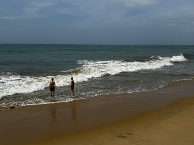 Zwei Personen stehen am Strand und beobachten die Wellen, während der Himmel leicht bewölkt ist und das Wasser glitzert.