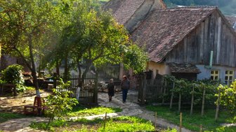 Zwei Personen gehen auf einem gepflasterten Weg durch einen malerischen Garten mit alten Häusern im Hintergrund.