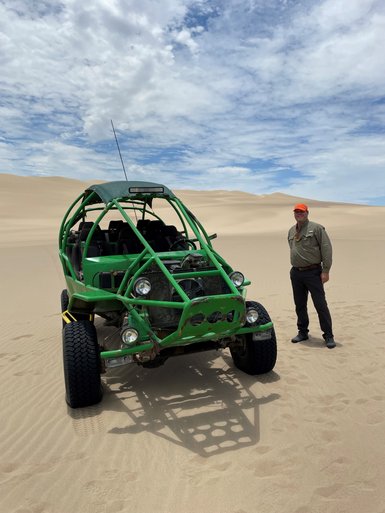 Ein Mann steht neben einem grünen Buggy in der Wüste, umgeben von sanften Sanddünen und einem bewölkten Himmel.