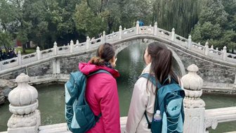 Zwei Frauen stehen auf einer Brücke und blicken auf einen ruhigen Teich, umgeben von üppigem Grün und historischen Gebäuden.