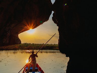 Ein Mann paddelt in einem roten Boot durch eine Höhle, während die Sonne hinter ihm aufgeht und den Himmel erleuchtet.