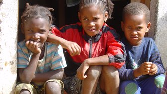Drei Kinder sitzen im Türrahmen eines Hauses, mit unterschiedlichen Gesichtsausdrücken, die den Moment lebendig machen.
