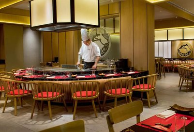 Ein Koch in einem eleganten Restaurant bereitet Speisen an einer zentralen Grillstation vor, umgeben von stilvollen Möbeln.