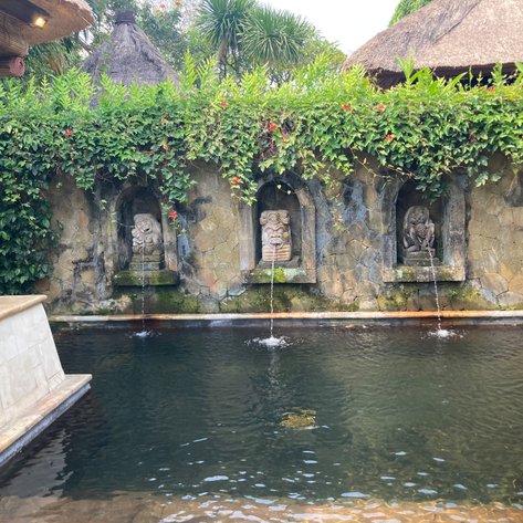 Ein ruhiger Pool mit drei kunstvollen Steinfiguren, aus denen Wasser sprudelt, umgeben von üppigem Grün und blühenden Pflanzen.