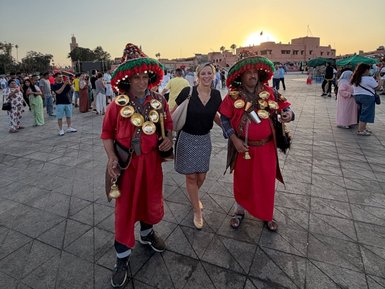 Zwei Männer in traditionellen roten Gewändern stehen zusammen mit einer lächelnden Frau vor einem belebten Platz in Marrakesch.