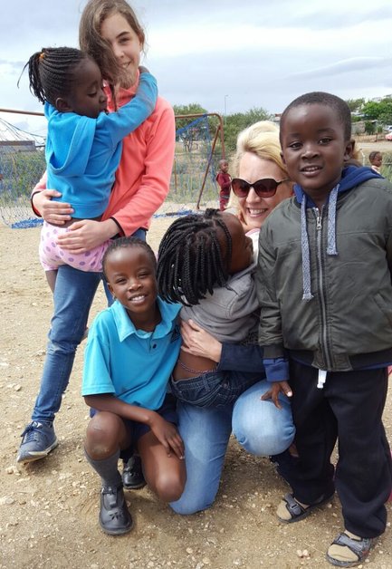 Eine Gruppe von Kindern und einer Frau lächelt fröhlich auf einem Spielplatz, umgeben von Sand und Spielgeräten.