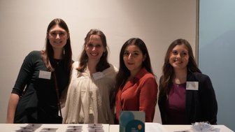 Vier Frauen stehen lächelnd an einem Empfangstisch, umgeben von Namensschildern und Informationsmaterialien.