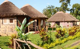 Drei traditionelle Hütten mit Strohdächern stehen in einem gepflegten Garten, umgeben von bunten Pflanzen und Bäumen.