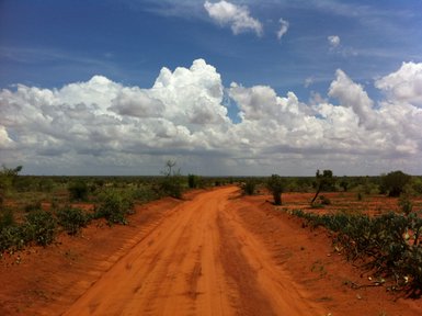 Eine staubige Straße schlängelt sich durch die weite, rote Landschaft des Tsavo Ost Nationalparks unter einem bewölkten Himmel.