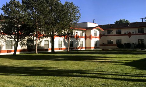 Ein helles Hotelgebäude mit orangefarbenen Akzenten, umgeben von gepflegtem Grün und hohen Bäumen im Vordergrund.
