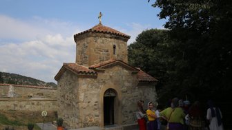 Eine kleine Kapelle aus Stein mit einem roten Ziegeldach und einem Kreuz, umgeben von Bäumen und Besuchern in traditioneller Kleidung.