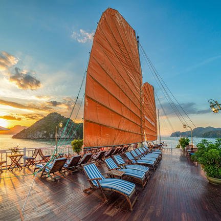 Die Terrasse eines Schiffes in der Ha Long Bucht mit orangefarbenen Segeln und Liegestühlen, während die Sonne untergeht.