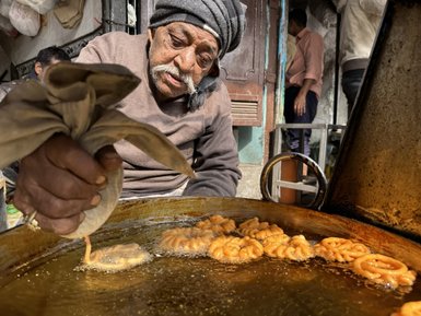 Ein älterer Mann bereitet frisch frittierte Süßigkeiten in einer großen Pfanne auf einem belebten Markt in Delhi zu.