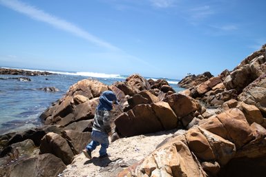 Entdeckungsfreudiges Baby geht vorsichtig über die Felsen an der Küste von Mossel Bay – Garden Route mit Kindern