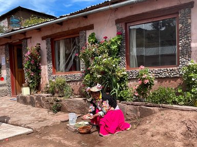Eine Frau in traditioneller Kleidung sitzt im Freien und wäscht Gemüse vor einem bunten, blühenden Garten.