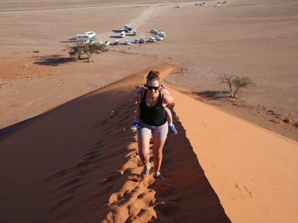Eine Mutter läuft mit Ihrem Kind auf dem Rücken eine Düne hinauf - Namibia Reise mit Kindern