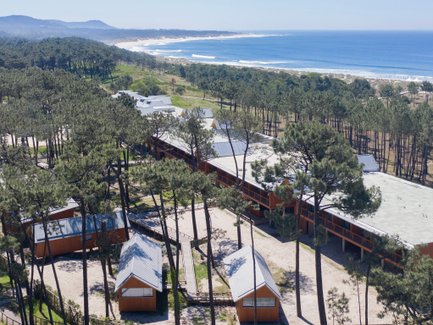 Eine Luftaufnahme zeigt ein modernes Hotel inmitten von Kiefern, mit Blick auf den Strand und das blaue Meer im Hintergrund.