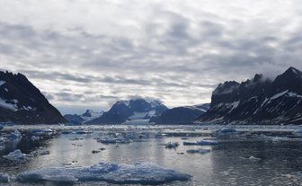 Eine ruhige, eisbedeckte Wasserfläche spiegelt die majestätischen Berge und den bewölkten Himmel in Hornsund wider.