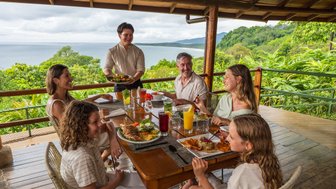 Eine Gruppe von sechs Personen genießt ein gemeinsames Essen auf einer Terrasse mit Blick auf das Meer und die grüne Landschaft.