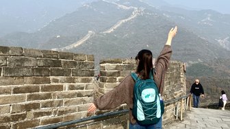 Eine Person mit einem Rucksack steht auf der Chinesischen Mauer und breitet die Arme in die Luft aus, um die Aussicht zu genießen.