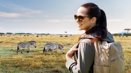 Eine lächelnde Frau mit Sonnenbrille steht in einer weiten Savanne, während Zebras im Hintergrund grasen.